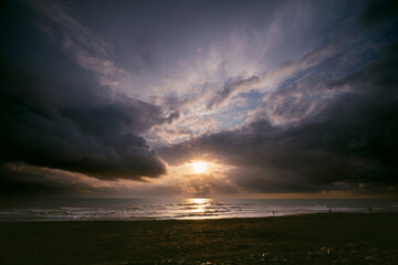 Sunset view, seashore, stormy waves, heavy rain clouds on horizon, sun breaking through the cloud...