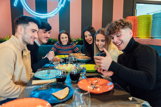 Positive young diverse friends sharing smartphone during dinner in restaurant