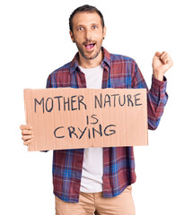 Young handsome man holding mother nature is crying protest cardboard banner screaming proud, celebrating victory and success very excited with raised arms