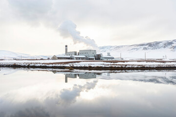 A geothermal power plant emits steam into the cold air, mirrored perfectly in the still waters below, with snow-capped mountains in the background