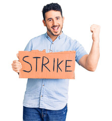 Young hispanic man holding strike banner cardboard screaming proud, celebrating victory and success very excited with raised arms