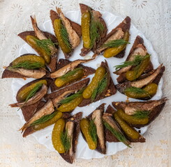 Sandwiches made of sprats, pickles and rye bread on a plate of white glass in close-up against a table covered with a light tablecloth