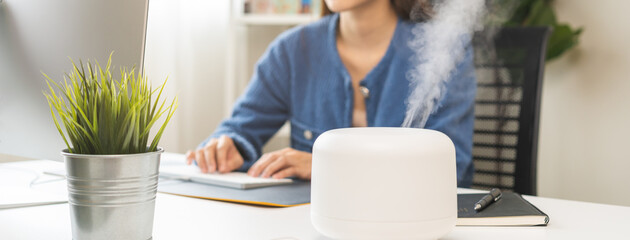 Modern air humidifier device during sitting workplace, happy asian young woman using computer work on internet, enjoying aromatherapy steam scent from essential oil diffuser in room at home office. - obrazy, fototapety, plakaty