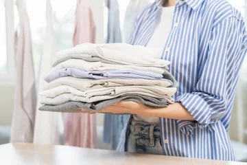 Fotobehang Feel softness, chore of pretty asian young woman hand holding pile clothing from table, stack folding clean clothes after washing, laundry and dry. Household working at home. Laundry of maid concept. © KMPZZZ