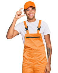 Young handsome african american man wearing handyman uniform smiling and confident gesturing with hand doing small size sign with fingers looking and the camera. measure concept.