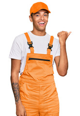 Young handsome african american man wearing handyman uniform smiling with happy face looking and pointing to the side with thumb up.