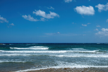 Fototapeta na wymiar Greek sandy beach. Ripple dark sea water, white foam, blue sky with cloud background, copy space.