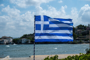 Greek flag on flagpole waving in the wind in front of blur island background. Greece sign symbol.