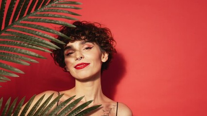 Fashion beauty portrait of young smiling brunette woman with evening stylish makeup and perfect clean skin. Sexy model with curly hair posing in studio. With red bright lips. Tropical palm leaf
