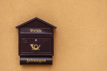 Brown mailbox with an emblem against a background of a plastered orange wall with gold lettering. Translation: "Letters", "Newspapers".