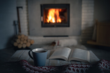 Cozy evening with reading near burning warm fireplace. Book and cup of tea on wooden table. Hygge...