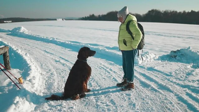 Happy beautiful 50s woman blowing snowflakes from her hands to her dog irish setter enjoying winter snow outdoors activity in winter day holiday. People and animals together having fun and smiling