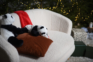 Christmas interior with a white sofa near green Christmas trees with lights that create a bokeh effect. two soft toy pandas with a red gnome hat on a white sofa.