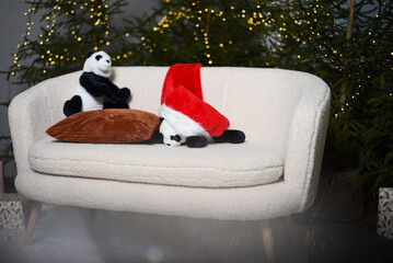 Christmas interior with a white sofa near green Christmas trees with lights that create a bokeh effect. two soft toy pandas with a red gnome hat on a white sofa.