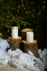 White LED candles that stand on fir tree stumps under fir trees with green needles in which Christmas lights are installed that create a bokeh effect.