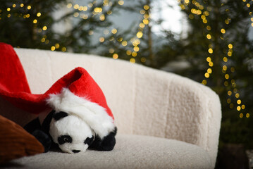 toy panda with a red gnome's Christmas hat is sitting on a white sofa near green fir trees in which the lights that create a bokeh effect are installed in the background