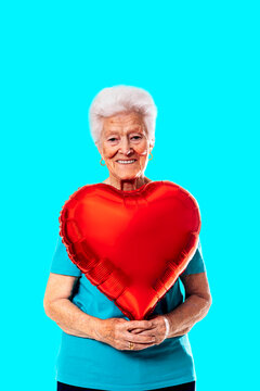 Smiling senior woman holding heart shaped balloon and looking at camera against blue background