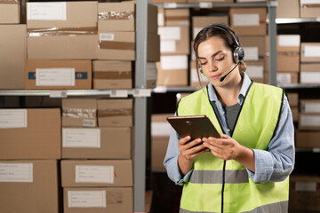 Female worker wearing headsets in logistics warehouse working with inventory app using digital...