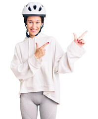 Beautiful brunette young woman wearing bike helmet and sporty clothes smiling and looking at the camera pointing with two hands and fingers to the side.