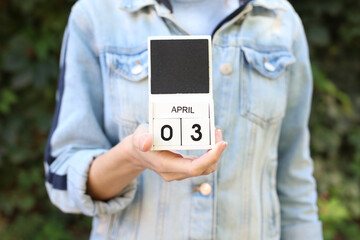 Woman in denim jacket holds white block wooden calendar with date April 3 outdoor