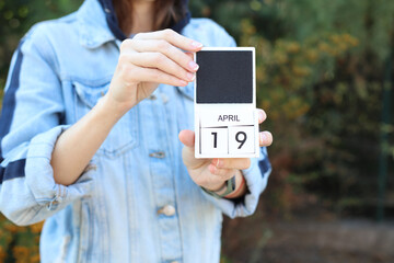 Woman in denim jacket holds white block wooden calendar with date April 19 outdoors