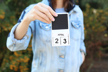 Woman in denim jacket holds white block wooden calendar with date April 23 outdoors