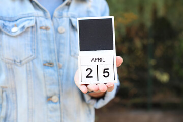 Woman in denim jacket holds white block wooden calendar with date April 25 outdoors