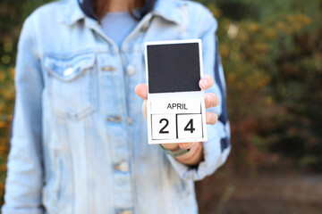 Woman in denim jacket holds white block wooden calendar with date April 24 outdoors
