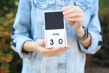 Woman in denim jacket holds white block wooden calendar with date April 30 outdoors