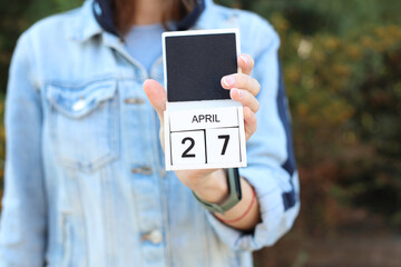 Woman in denim jacket holds white block wooden calendar with date April 27 outdoors