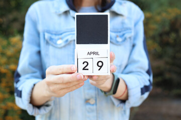 Woman in denim jacket holds white block wooden calendar with date April 29 outdoors