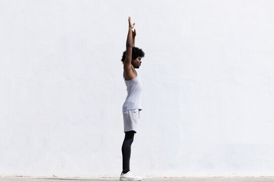 Full body side view of confident African American male athlete in activewear with concentration stretching arms while standing looking away against white background