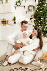 a young family with a small child is enjoying the new year at the Christmas tree in the kitchen or in a bright house, smiling, kissing and hugging congratulating each other celebrates the holiday