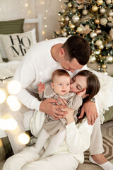 Obraz premium a young family with a small child is enjoying the New year at the Christmas tree on the bed, smiling, kissing and hugging congratulating each other celebrates the holiday