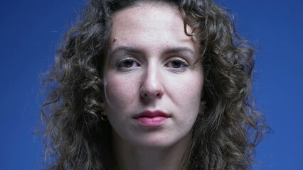 Close-up woman face looking at camera with neutral expression, blue background of person in 20s portrait