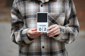 Woman in warm shirt holds calendar with date October 02 outdoors in autumn park