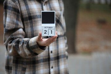 Woman in warm shirt holds calendar with date October 21 outdoors in autumn park
