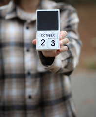 Woman in warm shirt holds calendar with date October 23 outdoors in autumn park