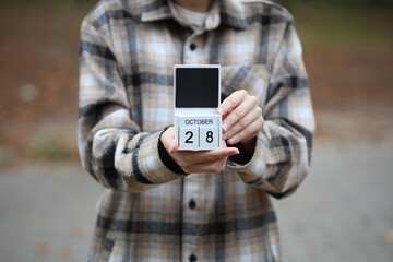Woman in warm shirt holds calendar with date October 28 outdoors in autumn park