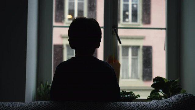 Silhouette of child staring at snow fall from home window during winter season. Back of young boy hypnotized by nature during moody day, December season