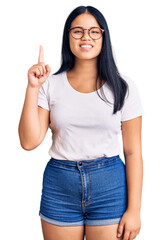 Young beautiful asian girl wearing casual clothes and glasses showing and pointing up with finger number one while smiling confident and happy.