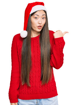 Young chinese woman wearing christmas hat surprised pointing with hand finger to the side, open mouth amazed expression.