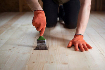 In the attic of the house, a young man varnishes the newly installed wooden floor with a brush so...