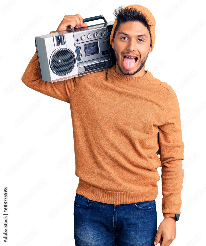 Wall mural Handsome latin american young man holding boombox, listening to music sticking tongue out happy with funny expression. emotion concept.