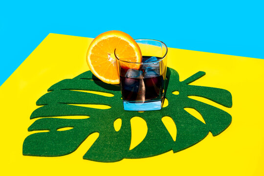 Cold beverage with slice of orange in glass on leaf decor over yellow backdrop against blue background in studio