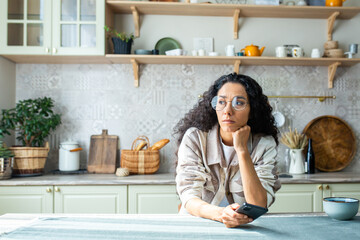 Lonely bored woman sitting alone at home in kitchen, hispanic woman with curly hair depressed thinking holding phone, got bad news message.