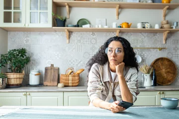 Foto op Plexiglas Lonely bored woman sitting alone at home in kitchen, hispanic woman with curly hair depressed thinking holding phone, got bad news message. © Liubomir