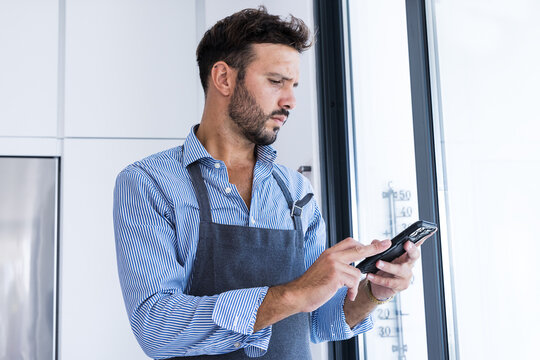 Side view of focused looking man in apron browsing smartphone in daylight near window
