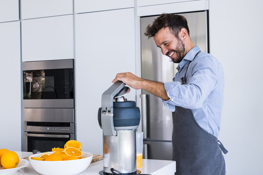 Happy adult man in apron standing near table with orange fruits squeezer in modern kitchen