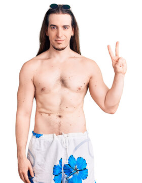 Young adult man with long hair wearing swimwear shirtless smiling with happy face winking at the camera doing victory sign. number two.